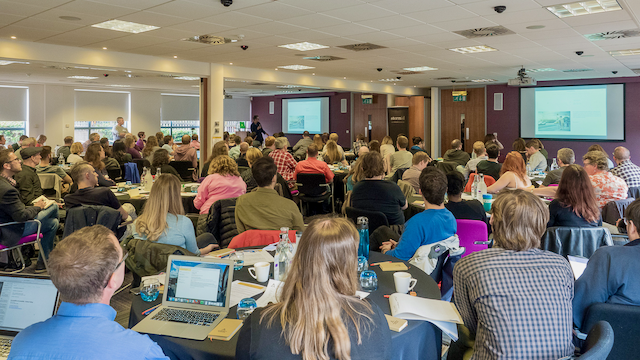 A packed conference room with attendees seated at tables, laptops open, watching a presentation